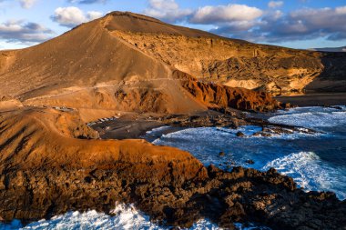 Charco Verde yakınlarındaki El Golfo 'daki Lanzarote' nin hava görüntüsü. Pas kayalıkları siyah lav kayalarıyla buluşur ve sörf yapar. Sahil yolu ve park halindeki araçlar öğleden sonra ışıklarıyla ziyaretçileri gösteriyor..