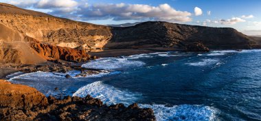Beyaz sörf pürüzlü lav kayalıklarına ve karanlık bir kumsal sahiline vururken pas kırmızısı uçurumlar ve bazalt tepeler Lanzarote 'de öğleden sonra ışıklarını yakalayarak erozyon ve lav akıntılarını gösteriyor..