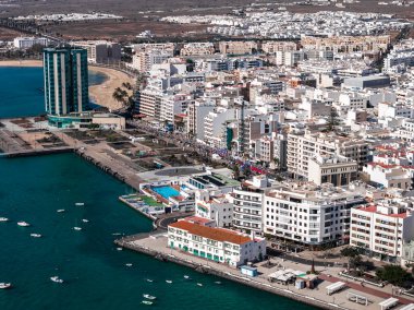Arrecife, Lanzarote 'nin hava görüntülerinde deniz kenarı gezinti alanı, küçük marina, Playa del Reducto ve Gran Hotel ve Spa görünüyor. Kalabalıklar ve pankartlar 2025 maraton güzergahını gösteriyor.