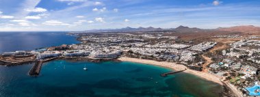 Lanzarote 'deki Costa Teguise hava manzarası turkuaz körfezler, altın kumlar, beyazlatılmış binalar, volkanik koniler, koruma altındaki koylar, bir yat limanı, havuzlar ve gündüz vakti sakin denizler gösteriyor..