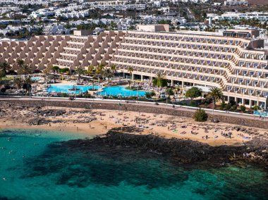Lanzarote, Kanarya Adaları 'ndaki teraslı bir otelin havadan görünüşü altın kum ve turkuaz Atlantik' e bakıyor, havuzlar, palmiyeler, gezinti güverteleri ve yüksek mevsimli kalabalıklar.