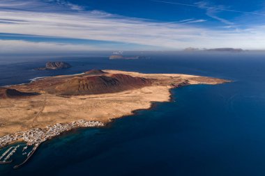 Kuzey Lanzarote açıklarındaki La Graciosa 'nın havadan görünüşü Montana Amarilla, Caldera de Agujas ve Caleta de Sebo marinasını turkuaz sığ, adacık, bulutlu ve kontrastlı gösteriyor..