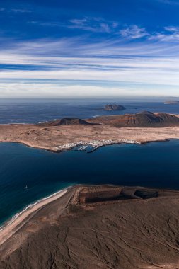 Hava sahnesi La Graciosa, Lanzarote ve Caleta de Sebo marinasını gösteriyor. Volkanik çorak topraklar, kumsallar, koniler, açık deniz adaları ve parlak gün ışığında katmanlı bulutlar..