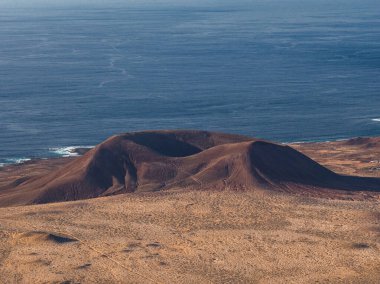 Havadan bakıldığında Lanzarote 'de Atlantik üzerinde lav alanları, toprak kül ovaları, soluk sörf ve gece yarısı aydınlatma sırtları şekillendiren pas kırmızısı bir volkanik koni ve krater görülüyor..