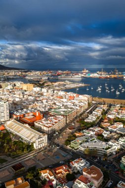 Hava görüntüsü Las Palmas de Gran Canaria limanı, Puerto de La Luz vinçleri, marina yelkenlileri, yapı gibi stadyum, kavşaklı sokaklar ve fırtına bulutları altında güneşli evler gösteriyor..