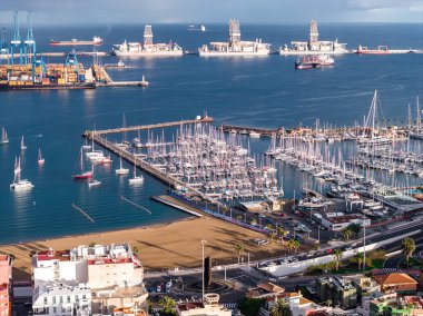 Las Palmas de Gran Canaria limanının hava görüntüsü marina direkleri, şehir plajı, mavi vinçler, kargo yığınları, sondaj gemileri ve kargo gemilerini öğleden sonra ışık altında gösteriyor..