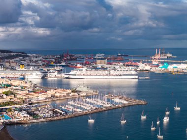 Las Palmas de Gran Canaria limanının hava görüntüsünde beyaz yolcu gemileri, kargo terminalleri, turuncu vinçler ve yelkenli, kış ışığı ve fırtına bulutları olan eğimli bir marina görülüyor..