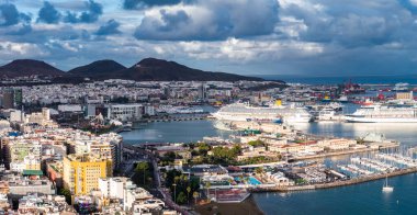 Las Palmas de Gran Canaria limanının hava görüntüsünde yolcu gemileri, marinalar, eğimli limanlar, sağa doğru vinçler ve yamalı bulutların altında Isleta 'nın karanlık ikiz tepeleri görülüyor..