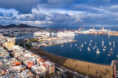 Hava görüntüsü Las Palmas de Gran Canaria limanı, Las Alcaravaneras plajı, beyaz yelkenli yatlar, yolcu gemileri, konteyner terminalleri ve kış ışığı altında şehir blokları gösteriyor..