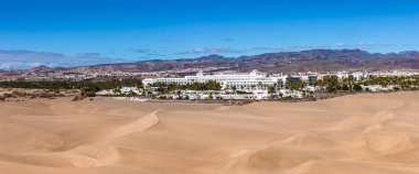 Geniş görüş, kayan altın Maspalom tepecikleri, kumda yürüyen aylaklar, palmiye güvertesi, beyaz sahil kenti, alçak şehir ve güney Gran Kanarya 'da berrak gökyüzü altında volkanik dağlar gösteriyor..