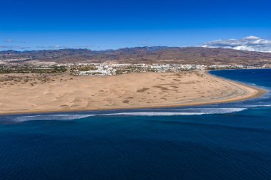 Maspaloma Kumulları 'nın havadan görünüşü, Gran Kanaryası' ndaki karanlık kum ve Atlantik. Rüzgar sırtları, çalılıklar, tatil köyleri, kurak dağlar, hafif dalgalar ve yürüyüşçüler görünür.
