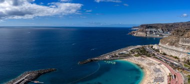 Gran Canaria 'daki Amadores Sahili' nin havadan görünüşü altın kum, kıvrımlı dalgalar, turkuaz bir lagün, tatil köyü uçurumları, kalabalık bir gezinti güvertesi, botlar ve yüzen bir su parkı gösteriyor..