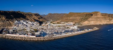 Hava görüntülerinde Puerto de Mogan, Gran Canaria, gemi direkleri ve yatları olan marina, beton tetrapodlar dalgalar, beyazlatılmış kanallar, palmiye sokakları, toprak kayalıklar, ayrılan küçük tekneler görülüyor..