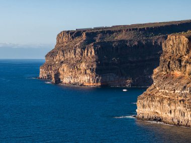 Puerto de Mogan yakınlarındaki Gran Canaria 'da Atlantik sularının üzerinde çizgili bazalt kayalıklar yükselirken, küçük beyaz bir tekne öğleden sonra sıcak ışıklarıyla üssü tarıyor..