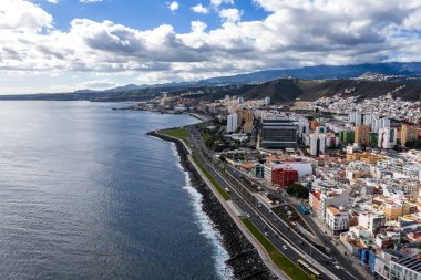 Las Palmas de Gran Canaria 'nın hava manzarası deniz kenarındaki karayolu, gezinti güvertesi, dalgakıran, cam ofis blokları, renkli evler, trafik, otobüsler ve volkanik dağları gösteriyor..