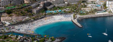 Gran Canaria 'daki Amadores Sahili' nin havadan görünüşü hilal kumu, taştan dalgalar, güneşlik, şemsiyeler, yüzücüler, botlar, gün ortasında bir katamaran ve teraslı oteller gösteriyor..