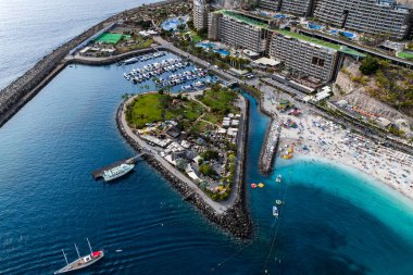 Hava görüntüsü, Anfi del Mar 'ın Gran Canaria' da kalp adası, bahçeler, plaj kulüpleri, beyaz yatlar, turkuaz sığlıklar, altın kum ve teraslı tatil beldeleri ile dolu bir marina olduğunu gösteriyor..