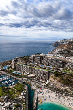Hava sahnesinde Amadores Sahili ve Anfi del Mar Gran Canaria 'da görülüyor. Kavisli dalgalar, basamaklı oteller, çatı havuzları, marina ve yumuşak ışık altında yoğun kumlu sahil..