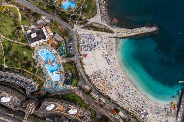 Hava görüntüsü, Gran Canaria 'daki Amadores Sahili' ni turkuaz körfezi, iki taş dalgası, parlak kumlu plaj, şemsiyeler, dinlenme yerleri, tatil köyü, göl havuzu ve öğle vakti gezinti alanı ile gösteriyor..