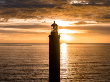 Maspalomas Deniz Feneri, Gran Canaria 'da gün batımında siluette duruyor. Dalgalar parlak bir yolu yansıtır, fener odası ve korkuluklar gevrek, sıcak bulutlar ufku çerçeveler..