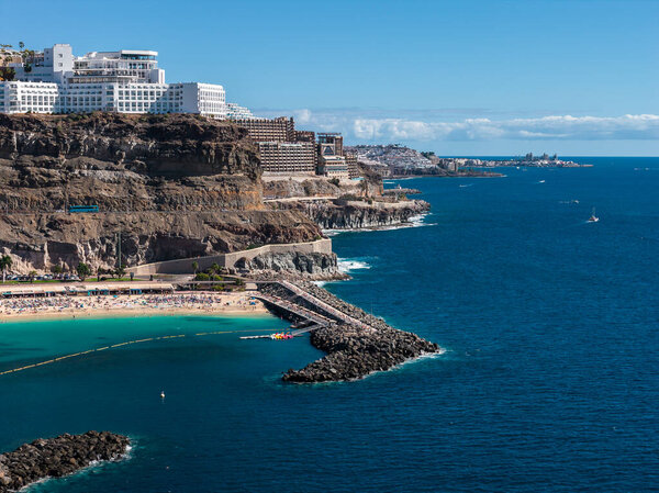Terraced resorts line cliffs above Amadores Beach in southwest Gran Canaria. Breakwaters shape a calm bay with sunbathers, boats on the Atlantic, and clear midday light.