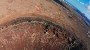 Fuerteventura 'nın volkanik iç mekânının hava görüntüsü krater jantları, lav yamaçları, seyrek çalılıklar, Atlantik ufku, toprak yolları, yürüyüşçüler ve gün ortası bulutları gösteriyor..