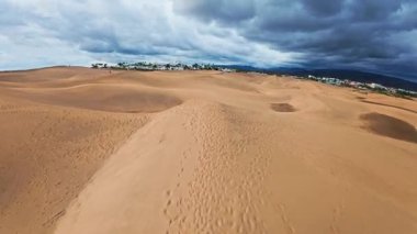 Maspaloma Kumulları, Büyük Kanarya üzerinde hava taraması. Drone kum tepeleri boyunca seyahat eder, ayak izleri ve görülebilir küçük figürler, palmiye ağaçları, beyaz tatil beldeleri, uzak dağlar.