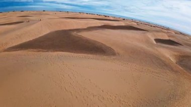 Hava aracı Maspalom kumulları üzerinde süzülüyor ve eğiliyor, Gran Canaria. Kıvrımlı sırtlar, volkanik ve bitki örtüleri, ziyaretçiler ve ayak izleri, ufukta beliren soluk Atlantik.