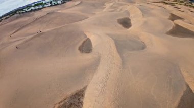 Hava aracı Gran Canaria 'daki Maspalom kumullarında süzülüyor, dalgalanan kumları, rüzgarın taradığı yamaları, ayak izlerini, küçük grupları ve fırtınalı bir gökyüzü altında tatil beldelerini gösteriyor..