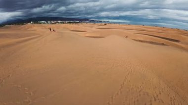 Gran Canaria 'daki Maspalom kumullarının hava görüntüsü kum tepeleri, tatil köyleri, palmiye ağaçları, yürüyen insanlar ve dağınık ışık bulutları altında Atlantik ufkunu gösteriyor..