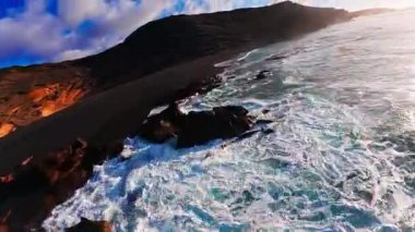 Lanzarote 'nin Los Hervideros ve El Golfo' daki hava manzarasında siyah kum, pas kayalıkları, pürüzlü kayalar, turkuaz sörfler, küçük bir lagün ve altın saat ışığı altında hareket eden eğim görülüyor..