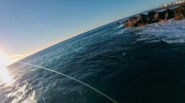 Hava görüntüsü, Lanzarote 'de siyah lav kayalıkları, çakıl taşı kıyıları ve Atlantik sörfü gösteriyor. Alçak altın güneş, yakınlarda beyazlatılmış evler ve yansımalar yaratır..