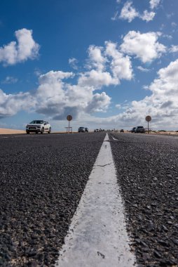 Büyük Kanarya 'daki kumullara doğru çatlak merkezin düşük açılı görüntüsü. Arabalar geçer ve gün ortası güneşinin ve bulutların altında kumlu bir sahil yolu boyunca dairesel işaretler durur..