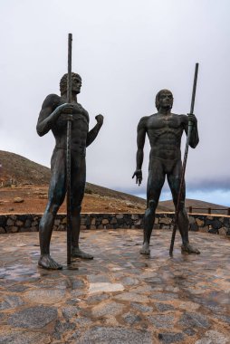 Mızraklı iki bronz Guanche savaşçı heykeli Betancuria yakınlarında duruyor, Fuerteventura, bulutlu gökyüzünün altında, İspanyol mirasını onurlandıran bir taş gözcü..