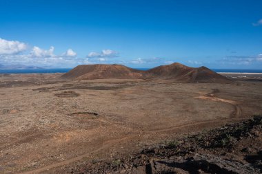 Timanfaya La Geria, Lanzarote 'de volkanik ovada iki yuvarlak kül konisi var. Nadir bitki örtüsü, koyu kahverengi arazi, öğlen ışığı, Atlantik ve kıyı kasabaları görülebilir..