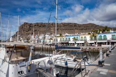 Yelkenliler, kalabalıklar palmiye ağaçlarının altında yürürken Puerto de Mogan rıhtımında kuyruk oluşturuyorlar. Parlak mavi ve yeşil süslemeli beyaz renkli binalar engebeli bir volkanik uçurumun altında otururlar..