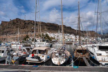 Yelkenliler ve yatlar Gran Canaria 'daki Puerto de Mogan marinasını dolduruyor. Beyazlatılmış binalar yamaçlara tırmanıyor, palmiye ağaçları yol alıyor, ve parlak bir gökyüzünün altında direkler yükseliyor..