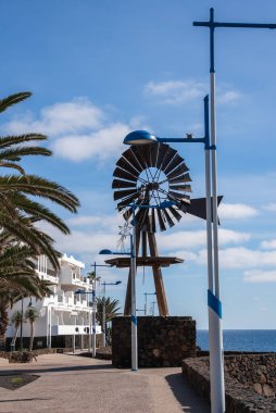 Metal bir yel değirmeni heykeli Lanzarote, Kanarya Adaları 'ndaki sahil geçidi boyunca bir taş üste duruyor. Palmiye ağaçları, beyaz binalar ve Atlantik açık gökyüzünün altında belirir..