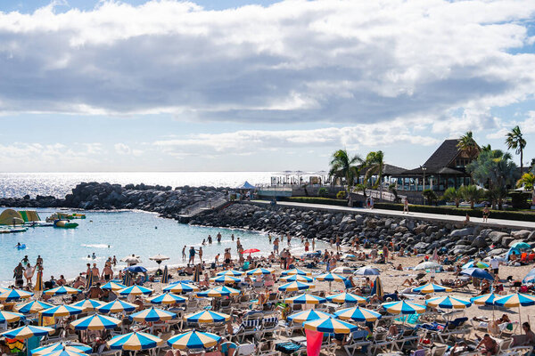 Rows of blue and white umbrellas fill golden sand by a cove on Gran Canaria. Palm lined promenade and beach bar sit above. People swim, kayak, and wade in calm water.