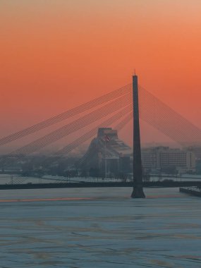 Vansu Köprüsü donmuş Daugava boyunca uzanırken kış günbatımı Riga, Letonya 'yı ısıtıyor. Letonya Ulusal Kütüphanesi arkasında yükseliyor, sis sürükleniyor, Letonya bayrağı dalgalanıyor.