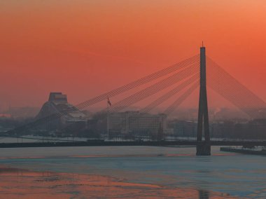 Vansu Köprüsü, solunda Letonya Ulusal Kütüphanesi ile Daugava Nehri boyunca uzanan buz tabakaları arasında yer alır. Sıcak turuncu ışık ve yumuşak sis lekesi..
