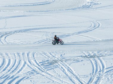 Hava görüntüsü, siyah kasklı ve turuncu yelekli bir motosikletlinin sert kar üzerinde kırmızı toprak bir bisiklet sürdüğünü ve parlak kış ışığı altında tekerlek izleri olduğunu gösteriyor..