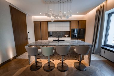 Çağdaş bir mutfak, içinde lavabo, dört bar taburesi, beyaz cam disk avizesi, sıcak ahşap kabin bulunan, ev aletleri ve taş kaplama ile mermer bir şelale adası sunar..