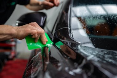 Bir teknisyen, su damlacıkları gibi boncuklar ve dikkatle vurgulanan sığ alan derinliği ile, bir kapı dikişi ve yan ayna yakınındaki berrak filmi pürüzsüz kılmak için parlak yeşil bir cam sileceği kullanır..