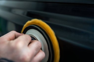 Detektörün elleri, koyu renk bir araba panelinde sarı köpük pedi olan bir cilalayıcıya rehberlik eder. Hareket, kenarı bulanıklaştırır. Yumuşak ışık girdapları ve yansımaları ortaya çıkarır..