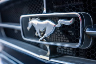 Yakın plan, klasik bir Ford Mustang ızgarasında krom koşu atı amblemini gösteriyor. Altıgen örgü, odağından yavaşça çıkmış, doğal ışık, yüzeysel alan derinliği..