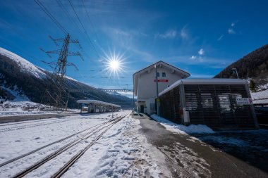 Küçük bir Zermatt alp istasyonu karlı dağlarda güneşlenir, uzun gölgeler düşer, ve paralel izler bir tünele çıkar, katenareri çizgileri ve kafes direkleri vardır..