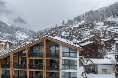 İsviçre, Zermatt 'ın hava panoramik görüntüsü dağ evi otellerini ve beyaz yamaçlardaki ahşap pansiyonları gösteriyor. Sisli dağ bulutlarının önünde modern cam bir balkon oteli duruyor..