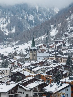 İsviçre, Zermatt 'ın karlı hava manzarası, kilise kulesi ve saati, dağ evi çatıları, koyu kozalaklı ormanlar, ve bulutların içinde Matterhorn ile sisli yamaçlar..