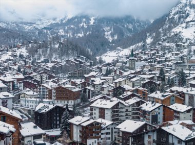 Zermatt, İsviçre 'nin havadan görünüşü, dağ evleri ve pansiyonlar üzerinde taze kar, kilise kulesi görünür, sis Matterhorn' u gizliyor, yumuşak ışık ve düşen tanecikler sessiz bir sabahı akla getiriyor.